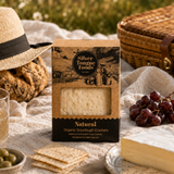 A box of Silver Tongue Foods Natural Organic Sourdough Crackers is styled on a picnic blanket with bread, cheese, grapes, olives, a drink, a straw hat, and a wicker picnic basket in an outdoor setting.