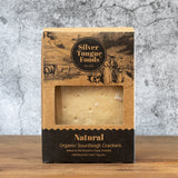 A package of Silver Tongue Foods Natural Organic Sourdough Crackers, shown in its brown kraft paper box with a clear window displaying the crackers inside, set against a textured gray background.